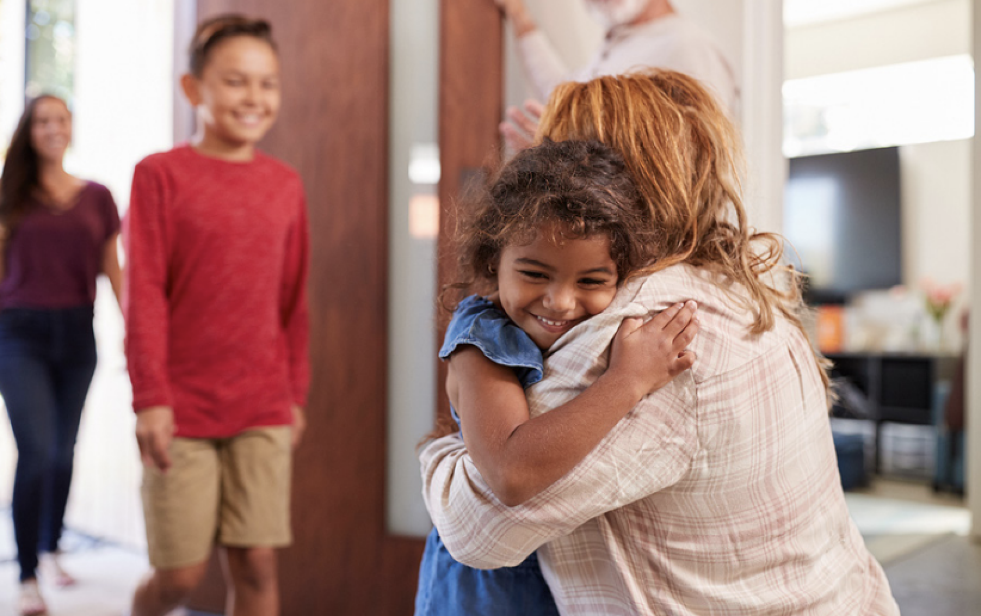 Young girl hugs a woman who waves at a boy and adult woman approaching.