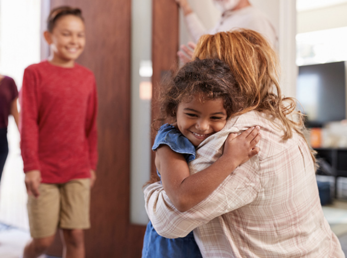 Young girl hugs a woman who waves at a boy and adult woman approaching.